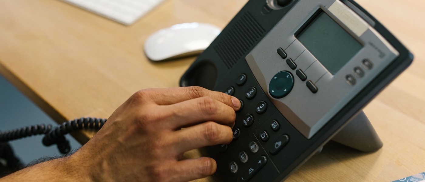 Main appuyant sur les touches d'un téléphone de bureau noir et gris posé sur un bureau en bois clair.