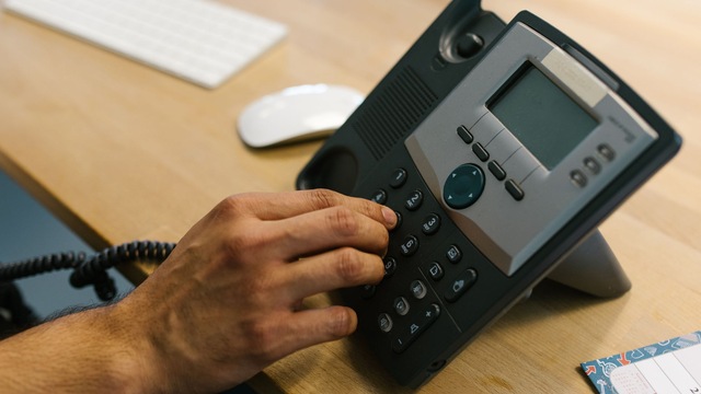 Main appuyant sur les touches d'un téléphone de bureau noir et gris posé sur un bureau en bois clair.