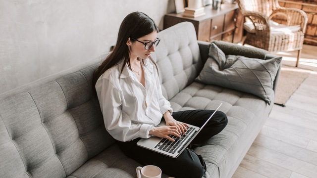 Femme assise sur un canapé gris, qui travaille sur un ordinateur portable.
